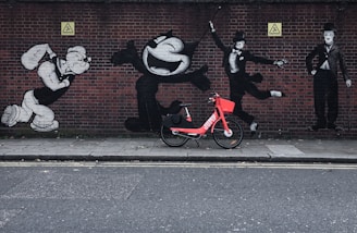 A vibrant red rental bike is parked in front of a brick wall adorned with black and white graffiti of famous cartoon and silent film characters in various dynamic poses.
