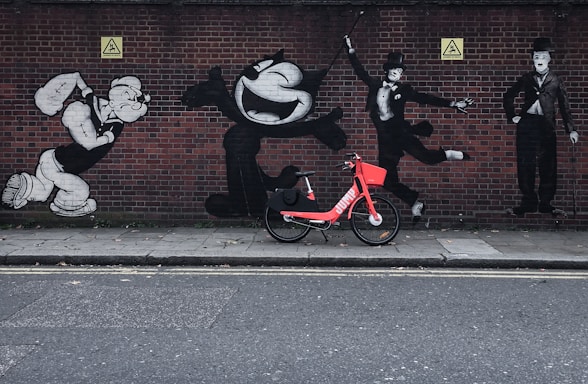 A vibrant red rental bike is parked in front of a brick wall adorned with black and white graffiti of famous cartoon and silent film characters in various dynamic poses.