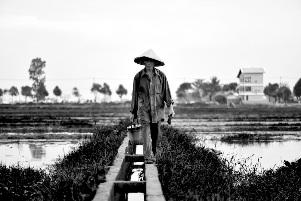 grayscale photography of man holding bucket while walking near rice field