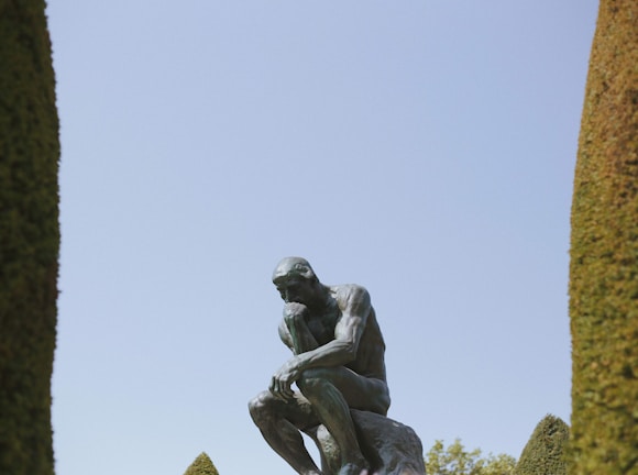 A bronze statue of a man deep in thought is positioned amid tall, manicured shrubs. The figure sits on a rock, chin resting on one hand, appearing contemplative under a clear sky.