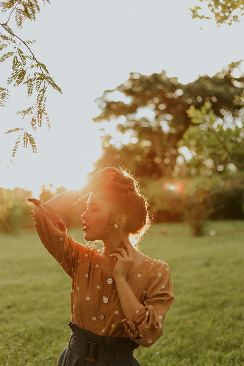 woman standing while touching her head and glancing right side on green field surrounded with tall and green trees during daytime