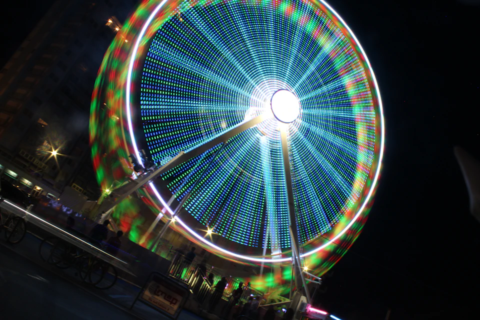 The image depicts a large, brightly lit Ferris wheel at night. The Ferris wheel is illuminated with blue and red lights, creating a vibrant contrast against the dark sky. The wheel is situated on a street corner, surrounded by buildings in the background. Several people can be seen standing around the base of the Ferris wheel, likely enjoying the spectacle or waiting to board it for their ride.