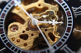 A close-up view of a mechanical watch's intricate inner workings, featuring gears and cogs in a polished gold tone. The watch hands and part of the dial with Roman numerals are also visible, adding elegance to the detailed craftsmanship.