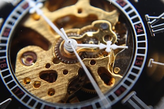 A close-up view of a mechanical watch's intricate inner workings, featuring gears and cogs in a polished gold tone. The watch hands and part of the dial with Roman numerals are also visible, adding elegance to the detailed craftsmanship.