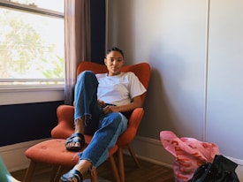 A person sits comfortably in a mid-century modern armchair with an orange upholstery. They are wearing a white t-shirt, blue jeans, and black sandals. The room is minimalistic with neutral-colored walls and hardwood flooring. A large window allows natural light to enter the room, casting shadows on the wall. There is a pink bag and a black bag placed on the floor nearby.