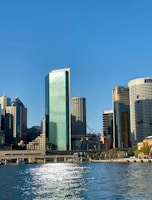 A cityscape featuring several skyscrapers with reflective glass facades under a clear blue sky. Sunlight glistens on the water of a body of water in the foreground. A crane is visible among the buildings, indicating ongoing construction.