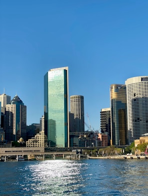 A cityscape featuring several skyscrapers with reflective glass facades under a clear blue sky. Sunlight glistens on the water of a body of water in the foreground. A crane is visible among the buildings, indicating ongoing construction.