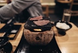 A table setting with a small grill on which slices of meat are being cooked. The grill rests on a stone base with written characters. Nearby, there is a bowl with white rice and condiments. The atmosphere feels intimate and cozy, suggesting a dining experience possibly in a restaurant.