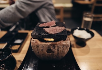 A table setting with a small grill on which slices of meat are being cooked. The grill rests on a stone base with written characters. Nearby, there is a bowl with white rice and condiments. The atmosphere feels intimate and cozy, suggesting a dining experience possibly in a restaurant.