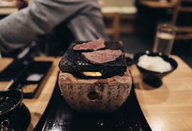 A table setting with a small grill on which slices of meat are being cooked. The grill rests on a stone base with written characters. Nearby, there is a bowl with white rice and condiments. The atmosphere feels intimate and cozy, suggesting a dining experience possibly in a restaurant.