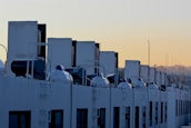 A row of solar water heaters is installed on the rooftop of a building, with the units aligned in parallel. The background features a clear sky transitioning into a warm, early morning or late afternoon glow.