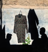 A dress with a floral pattern is hung on a hanger against a textured wall. The scene is shrouded in shadow, casting two distinct profiles on either side of the garment. A plant with broad leaves and a single pink flower grows in front.