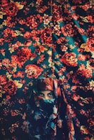 A person wearing a colorful, patterned headscarf is partially veiled against a background that also features a vivid floral design. The colors blend harmoniously with the backdrop, creating a seamless and artistic integration.