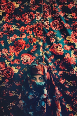 A person wearing a colorful, patterned headscarf is partially veiled against a background that also features a vivid floral design. The colors blend harmoniously with the backdrop, creating a seamless and artistic integration.