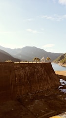 A large dam structure with a rocky surface dominates the foreground, leading towards a body of water. Trees and a small pavilion are situated on the top edge of the dam. Mountainous terrain spans the background under a clear sky, creating a serene landscape.
