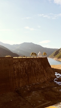 A large dam structure with a rocky surface dominates the foreground, leading towards a body of water. Trees and a small pavilion are situated on the top edge of the dam. Mountainous terrain spans the background under a clear sky, creating a serene landscape.