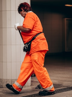 A person wearing bright orange work clothes is cleaning indoors, holding a mop. They have a fanny pack and are walking toward a tiled wall.