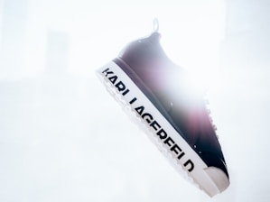 A single black and white sneaker is suspended in the air with the brand name 'KARL LAGERFELD' prominently displayed on the sole. The background is bright and overexposed, creating a high-contrast effect.