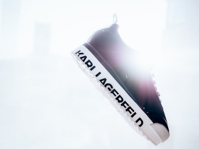 A single black and white sneaker is suspended in the air with the brand name 'KARL LAGERFELD' prominently displayed on the sole. The background is bright and overexposed, creating a high-contrast effect.