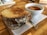 A grilled cheese sandwich with melty cheese between thick slices of toasted bread is placed on a wooden surface. Beside it, a cup filled with tomato soup sits on a white napkin with a silver spoon underneath.