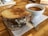 A grilled cheese sandwich with melty cheese between thick slices of toasted bread is placed on a wooden surface. Beside it, a cup filled with tomato soup sits on a white napkin with a silver spoon underneath.