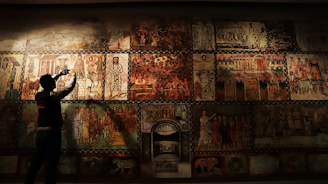 A person is standing in front of a large mural filled with intricate and colorful ancient-style paintings depicting various scenes and figures. The individual appears to be taking a photo with a smartphone, capturing the historical artwork displayed.