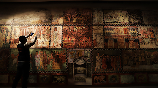 A person is standing in front of a large mural filled with intricate and colorful ancient-style paintings depicting various scenes and figures. The individual appears to be taking a photo with a smartphone, capturing the historical artwork displayed.