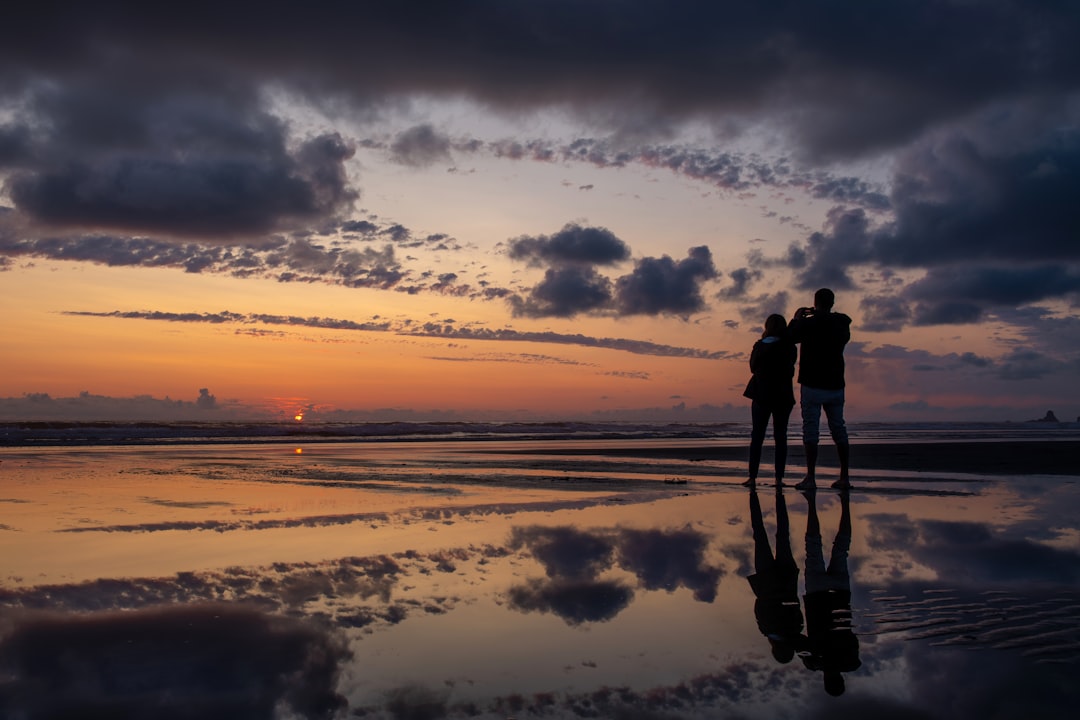 silhouette photography of man and woman standing on seashore during golden hour