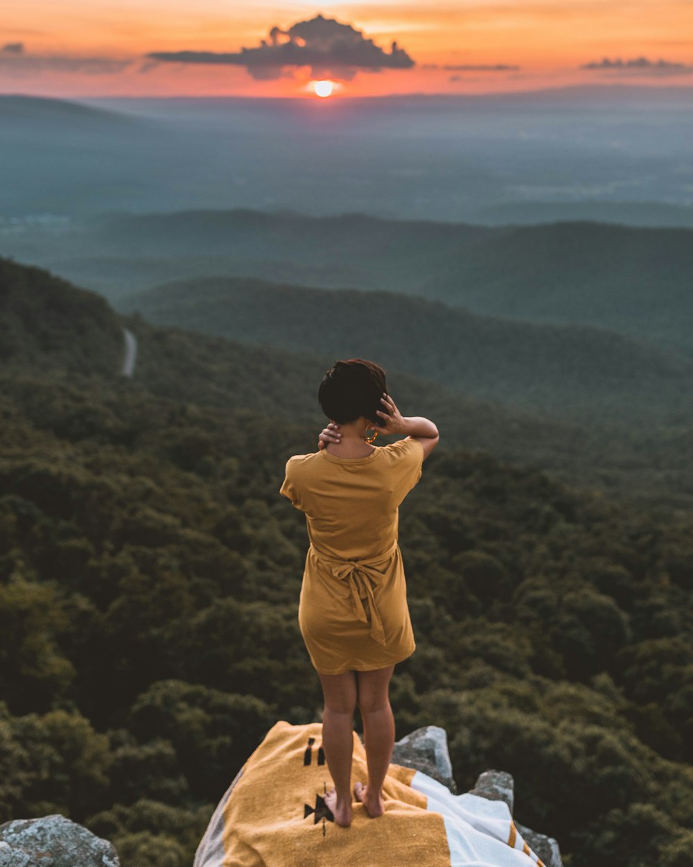 Frau im braunen Kleid steht während der goldenen Stunde auf einer Klippe mit Blick auf den Wald