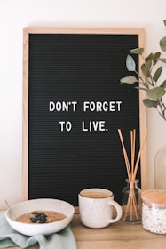 A black letter board with a wooden frame displays the message 'Don't forget to live.' Below the board, on a wooden surface, there is a bowl of oatmeal garnished with blueberries, a mug, a glass jar containing reeds, and a clear glass container. Some green leaves are also visible to the right of the board.
