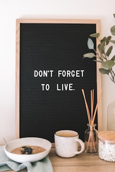 A black letter board with a wooden frame displays the message 'Don't forget to live.' Below the board, on a wooden surface, there is a bowl of oatmeal garnished with blueberries, a mug, a glass jar containing reeds, and a clear glass container. Some green leaves are also visible to the right of the board.