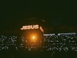 A dimly lit crowd is gathered in a large venue, with many holding up lights in the dark. The word 'JESUS' is prominently displayed in large, illuminated letters on a screen above the crowd, with a bright, warm light shining from the center.