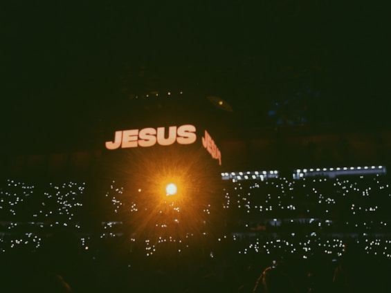 A dimly lit crowd is gathered in a large venue, with many holding up lights in the dark. The word 'JESUS' is prominently displayed in large, illuminated letters on a screen above the crowd, with a bright, warm light shining from the center.