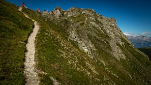 A narrow hiking trail winds up a grassy, rocky mountain slope. Several hikers are visible near the top, enjoying the sunny weather and expansive mountain views in the background.