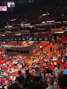 An indoor sports arena filled with a large crowd of spectators. People are sitting in tiered seating areas with red and orange tones dominating the seats. A person in the foreground is waving a large white flag with 'MIAMI' written on it. Numerous attendees wear basketball jerseys, and there is visible excitement and energy in the atmosphere. Advertisements for Uber are displayed prominently on digital boards near the ceiling.