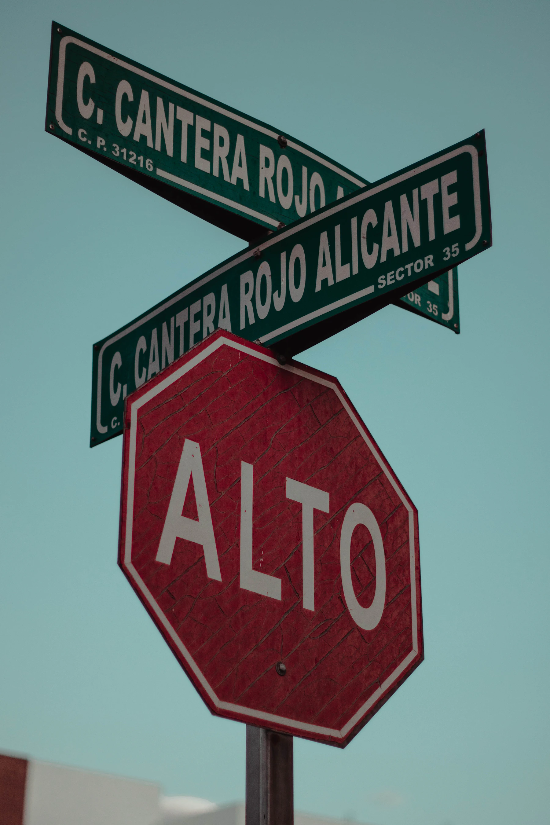 A red octagonal stop sign reading ALTO under two green Mexican street-name signs, against a teal sky