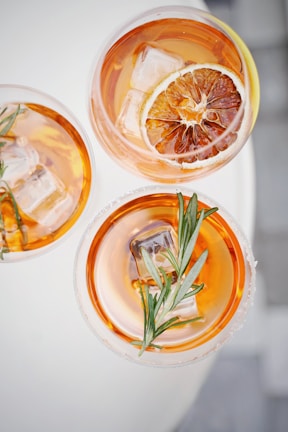 Three glasses of an amber-colored cocktail are garnished with sprigs of rosemary and a slice of dried citrus. The drinks contain ice cubes and have a sophisticated presentation against a light background.