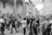 A group of people, many with cameras and phones, are gathered in an outdoor setting, possibly a historic or tourist site. They appear to be taking photos and looking at something off-camera, surrounded by classic architecture and statues.