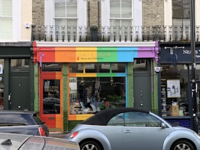 A storefront with a vibrant, colorful facade featuring red, orange, green, blue, and purple panels. The shop sign mentions a living and giving shop for Save the Children. In front, a light blue convertible car is parked. The shop windows display clothes and other items.