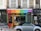A storefront with a vibrant, colorful facade featuring red, orange, green, blue, and purple panels. The shop sign mentions a living and giving shop for Save the Children. In front, a light blue convertible car is parked. The shop windows display clothes and other items.
