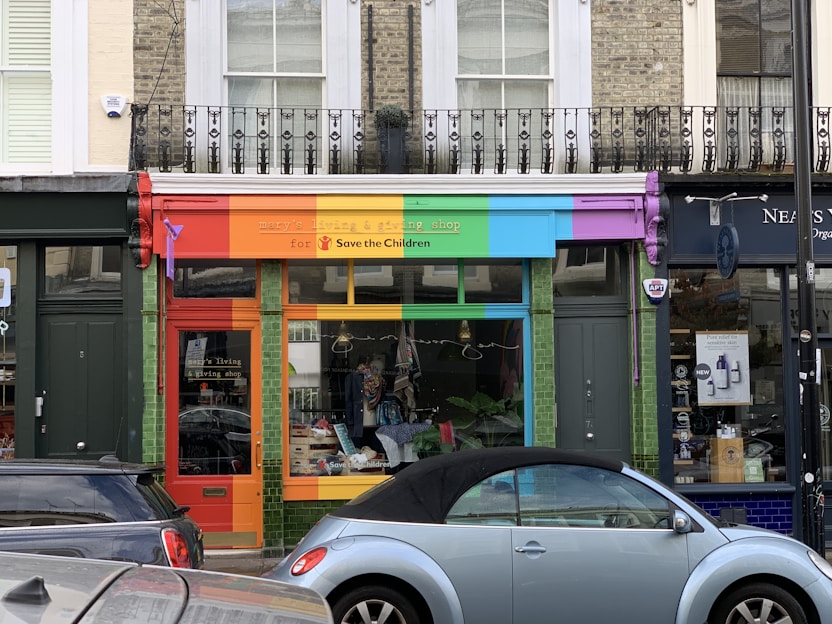 A storefront with a vibrant, colorful facade featuring red, orange, green, blue, and purple panels. The shop sign mentions a living and giving shop for Save the Children. In front, a light blue convertible car is parked. The shop windows display clothes and other items.