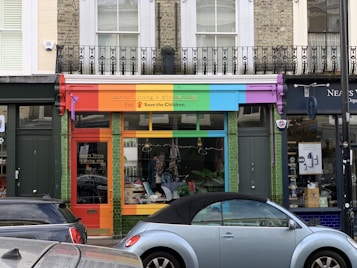 A storefront with a vibrant, colorful facade featuring red, orange, green, blue, and purple panels. The shop sign mentions a living and giving shop for Save the Children. In front, a light blue convertible car is parked. The shop windows display clothes and other items.