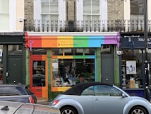 A storefront with a vibrant, colorful facade featuring red, orange, green, blue, and purple panels. The shop sign mentions a living and giving shop for Save the Children. In front, a light blue convertible car is parked. The shop windows display clothes and other items.