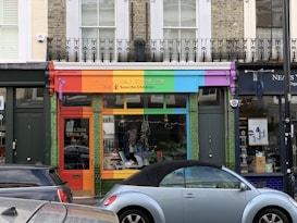 A storefront with a vibrant, colorful facade featuring red, orange, green, blue, and purple panels. The shop sign mentions a living and giving shop for Save the Children. In front, a light blue convertible car is parked. The shop windows display clothes and other items.