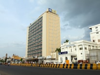 A large, multi-story building with a sign displaying 'LIC' at the top dominates the skyline. Next to it, there is a white colonial-style building with 'State Bank of India' signage. The scene includes a wide road in the foreground with yellow and black traffic barriers, a few vehicles and pedestrians. The sky is partly cloudy, suggesting changing weather conditions.