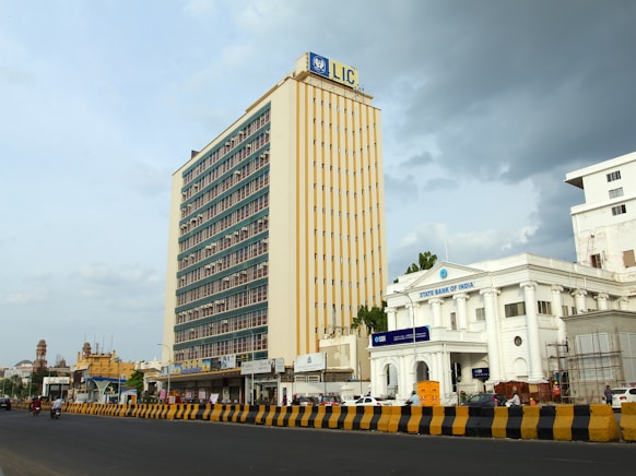 A large, multi-story building with a sign displaying 'LIC' at the top dominates the skyline. Next to it, there is a white colonial-style building with 'State Bank of India' signage. The scene includes a wide road in the foreground with yellow and black traffic barriers, a few vehicles and pedestrians. The sky is partly cloudy, suggesting changing weather conditions.