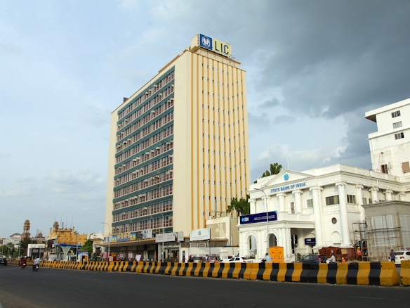 A large, multi-story building with a sign displaying 'LIC' at the top dominates the skyline. Next to it, there is a white colonial-style building with 'State Bank of India' signage. The scene includes a wide road in the foreground with yellow and black traffic barriers, a few vehicles and pedestrians. The sky is partly cloudy, suggesting changing weather conditions.