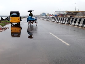 A wet road with reflections of a yellow auto rickshaw and a person on a cycle rickshaw holding an umbrella. The road is bordered by a black and white striped barrier, and in the background, there are buildings and some greenery. The overall scene suggests recent rainfall.