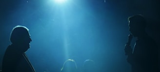 Two silhouetted figures are facing each other in a dimly lit space, with one holding a microphone. A bright blue light illuminates the scene from above, creating a dramatic atmosphere with visible beams extending downward.