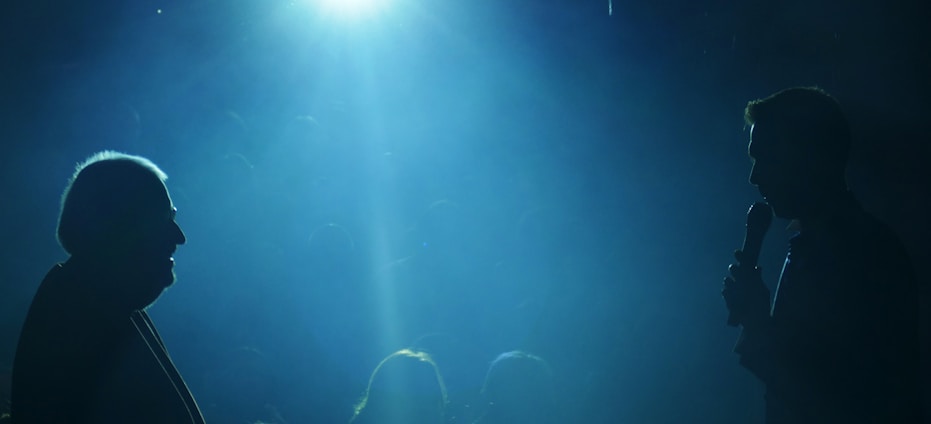 Two silhouetted figures are facing each other in a dimly lit space, with one holding a microphone. A bright blue light illuminates the scene from above, creating a dramatic atmosphere with visible beams extending downward.